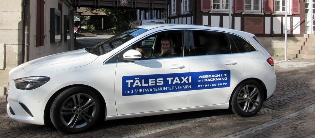 Eine weiße Mercedes B-Klasse von Täles Taxi mit blauer Aufschrift vor Fachwerkhäusern