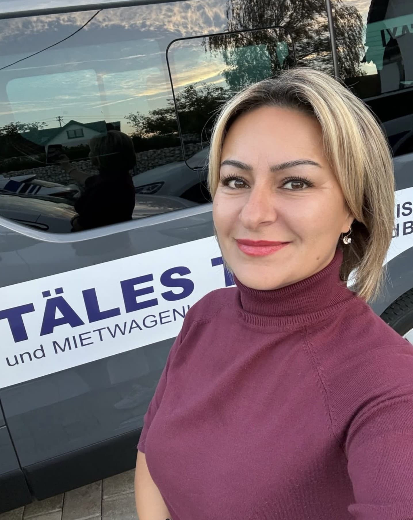 Pembe Akgün, lächelnd, vor einem Wagen von Täles Taxi mit der blauen Aufschrift „Täles Taxi und Mietwagen“